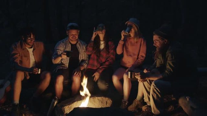 无忧无虑的游客烘烤着碰杯，然后晚上在森林里坐在火堆旁喝酒