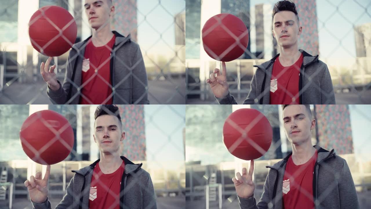 一位英俊的年轻足球运动员看着相机，手指上旋转着一个红色足球的肖像。时尚的足球运动员站在城市环境中的篱