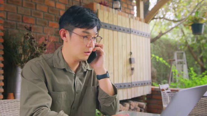 亚洲自由职业者戴着眼镜在电话上聊天，使用笔记本电脑。多任务和远程工作概念。