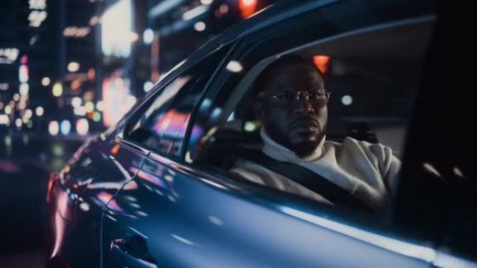 时髦的戴眼镜的黑人在晚上坐出租车的后座上下班。英俊的男性乘客在城市街道上开着霓虹灯的汽车时看着窗外。