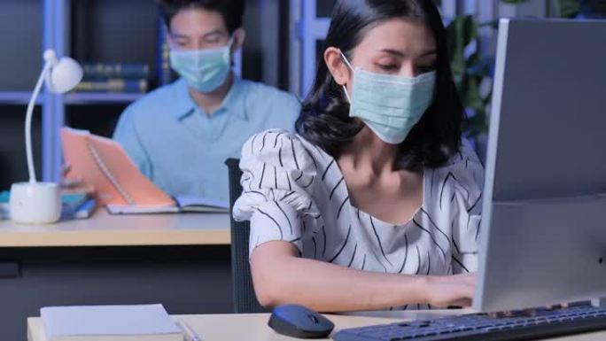 新型冠状病毒肺炎大流行后，在办公室里戴着电脑的亚洲女商人戴着口罩，而同事在晚上工作到很晚。在 “新”