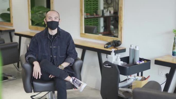 发廊的亚洲华人发型师老板看着镜头，带着面具自信地微笑