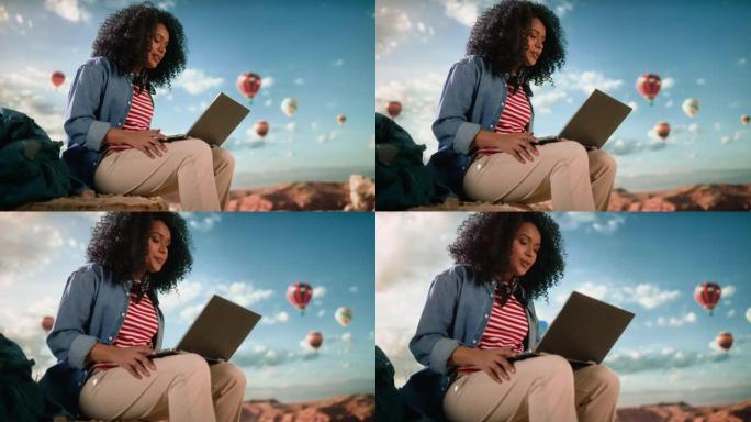 激动的美丽的年轻多民族女性旅行者，非洲发型，在落基峡谷山谷顶部的笔记本电脑上通过视频通话交谈。热气球
