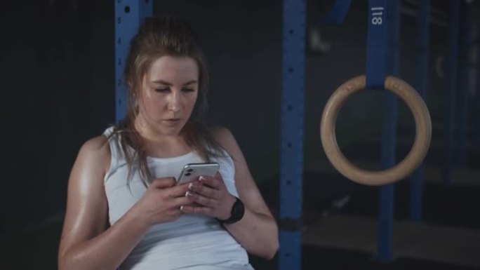美丽的高加索人适合出汗的年轻女子使用智能手机健身应用程序在现代健身房锻炼后跟踪活动。