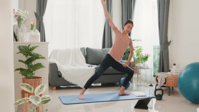年轻迷人的亚洲女性穿着运动服，在平板电脑上观看健身在线视频，为初学者在家里的客厅练习瑜伽。
