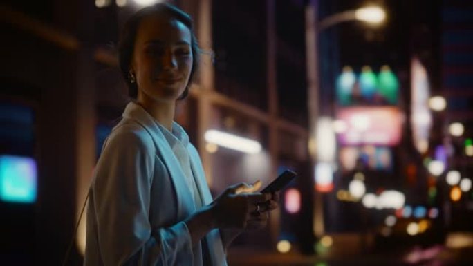 美丽的年轻女子使用智能手机走过充满霓虹灯的夜晚城市街道。微笑的女性使用手机进行在线购物，社交媒体发布