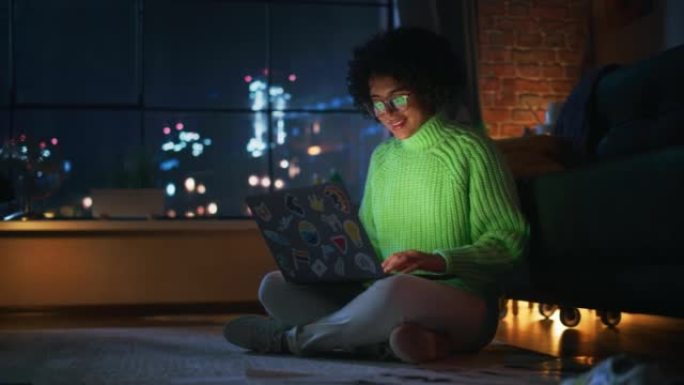 积极的拉丁女性在家里使用带贴纸的笔记本电脑。女人躺在地板上，微笑着在电脑上发短信。窗户后面的夜景