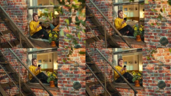 乐观，微笑的穆斯林女性坐在舒适的褐石屋公寓的窗台上使用智能手机。查看社交媒体的女孩。从楼外向下倾斜拍