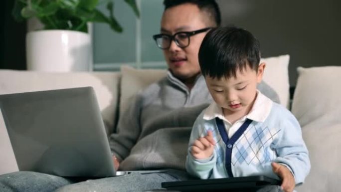 父亲使用笔记本电脑，儿子在沙发上使用平板电脑