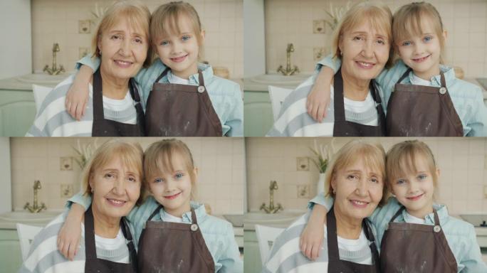 小女孩和成熟女人穿着围裙站在厨房拥抱并看着相机的肖像