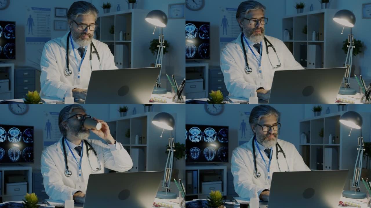 成熟的医生晚上在黑暗的办公室里用笔记本电脑工作，喝咖啡