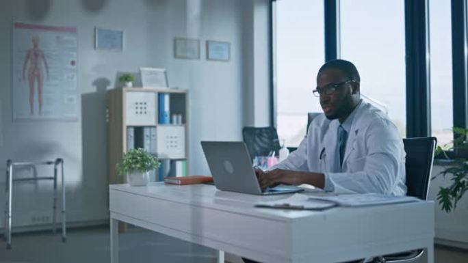 戴眼镜的冷静的非洲裔美国家庭医生正在一家健康诊所的笔记本电脑上工作。穿着白大褂的医生正在医院办公室的