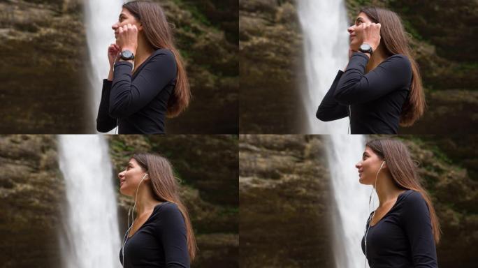 微笑的年轻女子站在瀑布前插入入耳式耳机，在智能手机上听音乐