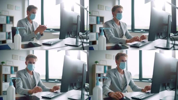 戴着防护口罩的英俊时尚年轻人在办公室的电脑上在办公桌前工作时使用洗手液。蓝眼睛金发年轻男性遵循健康和