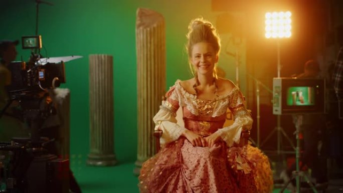 微笑的女演员穿着文艺复兴时期的连衣裙，坐在椅子上看着相机。背景中的绿色屏幕。工作室拍摄历史大片。论电