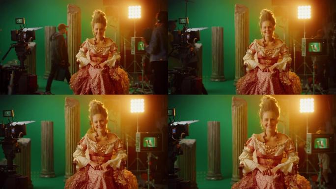 微笑的女演员穿着文艺复兴时期的连衣裙，坐在椅子上看着相机。背景中的绿色屏幕。工作室拍摄历史大片。论电