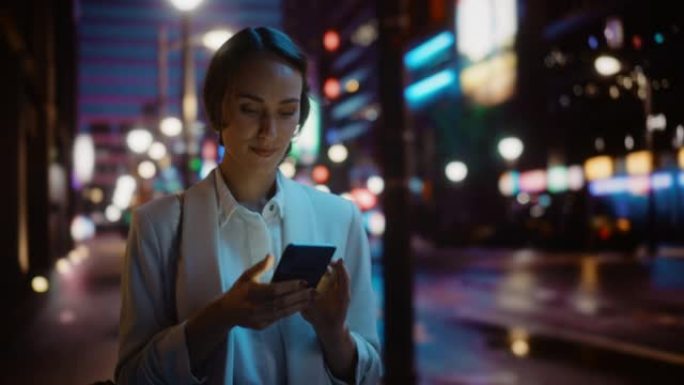 美丽的年轻女子使用智能手机走过充满霓虹灯的夜晚城市街道。使用手机，发布社交媒体，在线购物，发短信的华