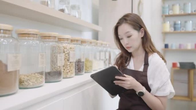 36岁的漂亮亚洲年轻女子的肖像，穿着棕色围裙，用平板电脑工作，同时检查库存在可持续的零浪费杂货店。可