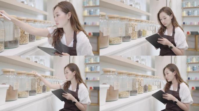36岁的漂亮亚洲年轻女子的肖像，穿着棕色围裙，用平板电脑工作，同时检查库存在可持续的零浪费杂货店。可