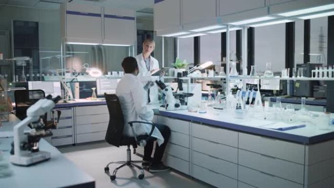 女性和男性医学研究科学家在显微镜下进行实验时进行对话，在平板电脑上写下分析结果。现代生物科学实验室。