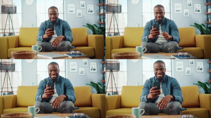 兴奋的黑人非洲裔美国人在客厅的沙发上休息时使用智能手机。快乐的男人在家里微笑，通过互联网与同事和客户