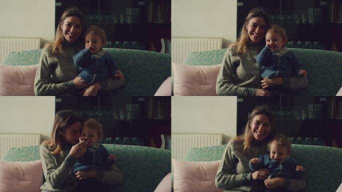 一个年轻快乐的母亲和她蹒跚学步的男婴坐在家里客厅的沙发上，对着镜头微笑。幸福的概念，童年，生活，母性