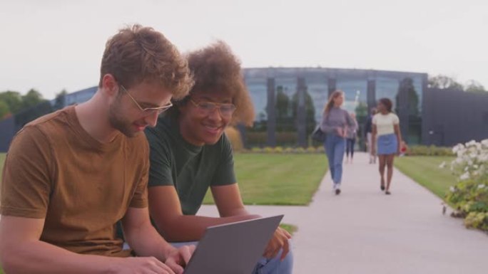 男性大学或大学生坐在户外的校园里聊天并在笔记本电脑上工作