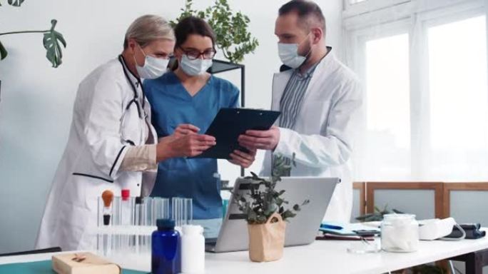 诊所的团队合作。三名穿着实验服和磨砂膏的医生讨论案例，看看现代轻型医院办公室的笔记本电脑。
