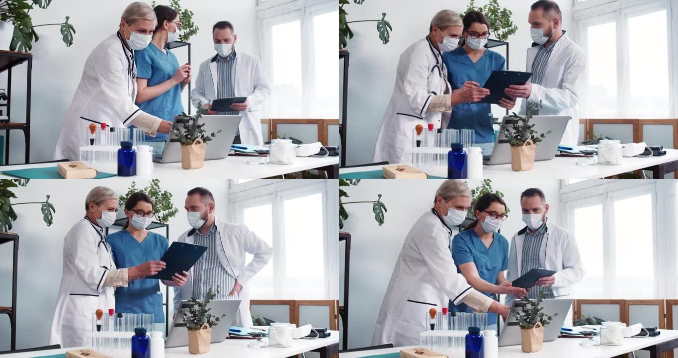 诊所的团队合作。三名穿着实验服和磨砂膏的医生讨论案例，看看现代轻型医院办公室的笔记本电脑。