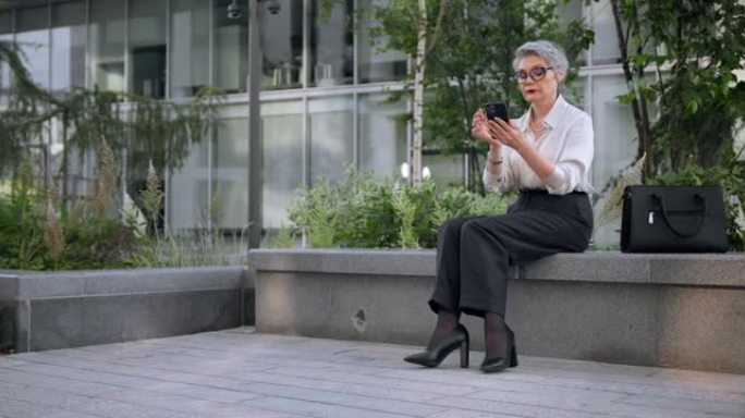一位穿着黑色裤子和时尚高跟鞋的白发漂亮商务女士正坐在商务中心前的石凳上，在手机里发短信