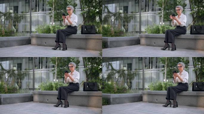 一位穿着黑色裤子和时尚高跟鞋的白发漂亮商务女士正坐在商务中心前的石凳上，在手机里发短信