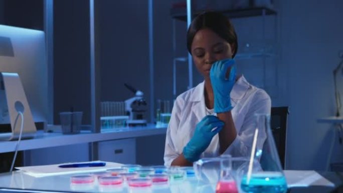 一位年轻的黑皮肤微生物学家戴着防护眼镜，手套和白色实验室外套，开始在生物危害实验室的培养皿上观察结果