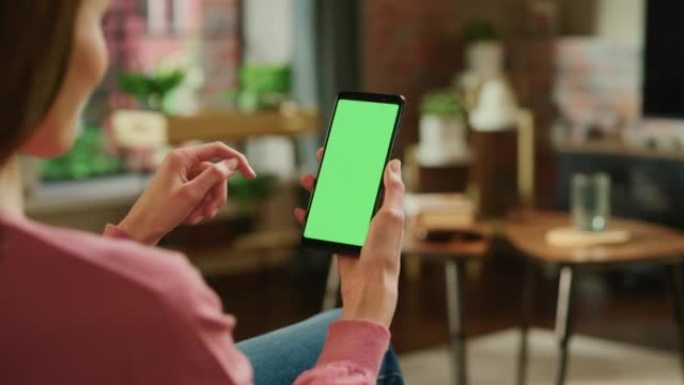 带有绿屏模拟显示的智能手机上的女性手滚动提要。女性正在家里休息，并在移动设备上查看社交媒体。特写镜头