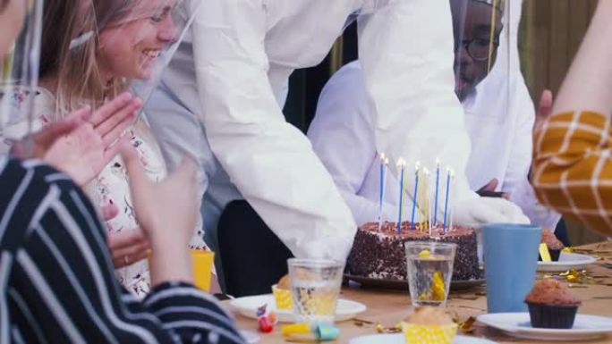年轻快乐的金发美女和朋友们一起庆祝生日，戴着COVID-19塑料面罩吹蛋糕