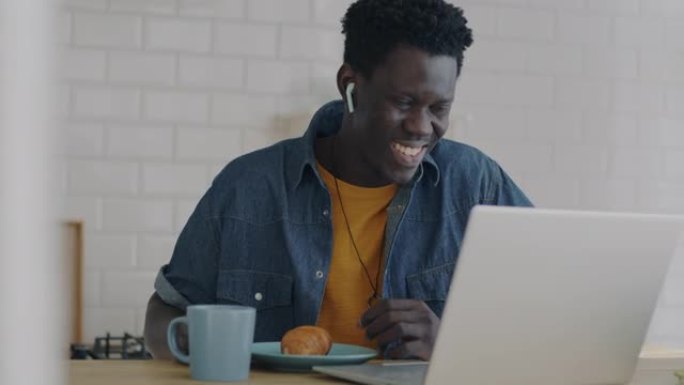 快乐的非洲裔美国人吃羊角面包，笑着看着家里厨房里的笔记本电脑屏幕
