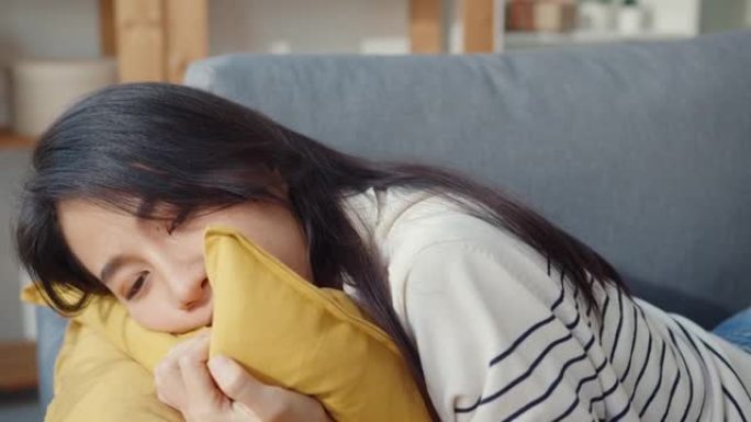 年轻的亚洲妇女躺在客厅舒适的沙发上，每天辛苦工作后睡觉，感到恶心和不好，疲倦的女性在家中躺在沙发上入