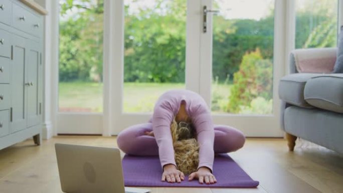年轻女子坐在家里的垫子上，拿着笔记本电脑在网上上瑜伽课