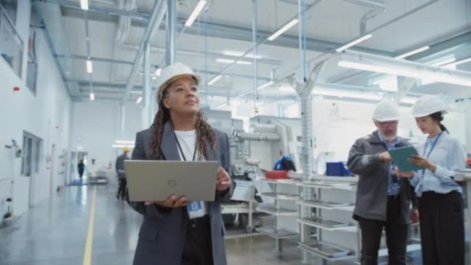 在电子制造工厂，一位戴着安全帽的黑人女工程师的肖像在使用笔记本电脑。处理日常任务和研发数据的技术人员