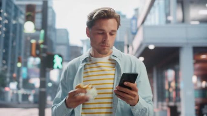 一个英俊的年轻人穿着便服，使用智能手机并在街上吃汉堡的肖像。大城市的经理与人们在线联系，消息传递和浏