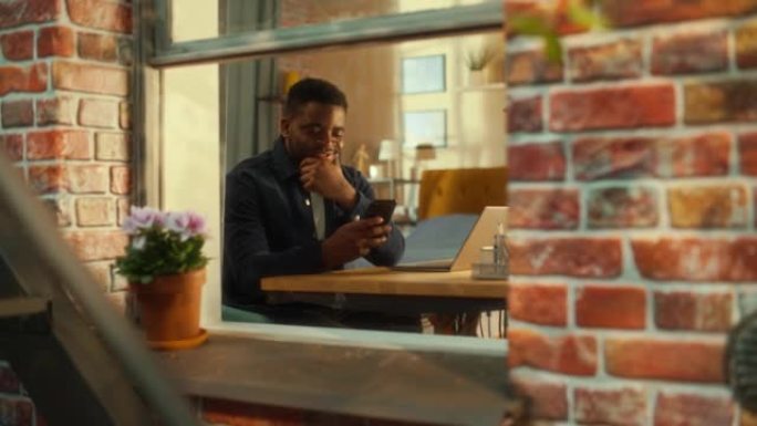 快乐的年轻黑人男性坐在家里，在社交媒体上与朋友联系，使用智能手机上的在线应用程序。自由职业者在他时尚