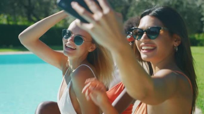 穿着泳衣的墨镜的年轻多民族女友的慢动作特写，他们很高兴一起享受暑假，并在阳光明媚的日子在游泳池里自拍