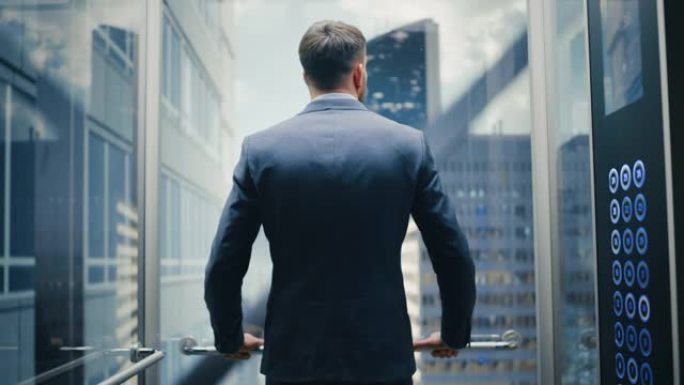 成功的商人穿着西装骑着玻璃电梯到现代商务中心的办公室。年轻的男性在电梯的全景窗口外看着现代的市中心摩