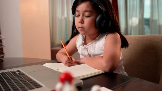 亚洲女孩在网上学习期间通过笔记本电脑和耳机做笔记，笔记本电脑在前台。社交距离，儿童教育，自学，电子学