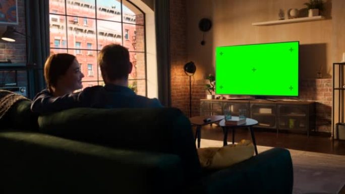 年轻的夫妇花时间在家里，坐在沙发上，在他们时尚的阁楼公寓里看绿屏模拟显示的电视。男人和女人流媒体电影