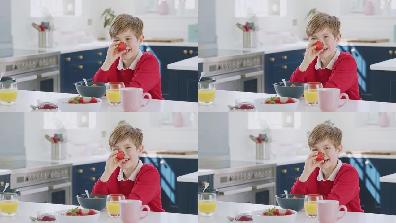 笑着的男孩穿着校服在厨房里吃早餐时把草莓放在鼻子上