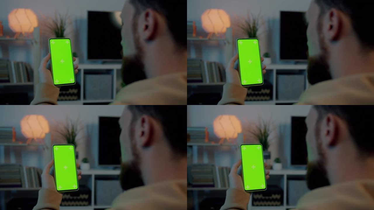 男性手持色度键绿屏智能手机的特写镜头，男性晚上在家观看显示的内容