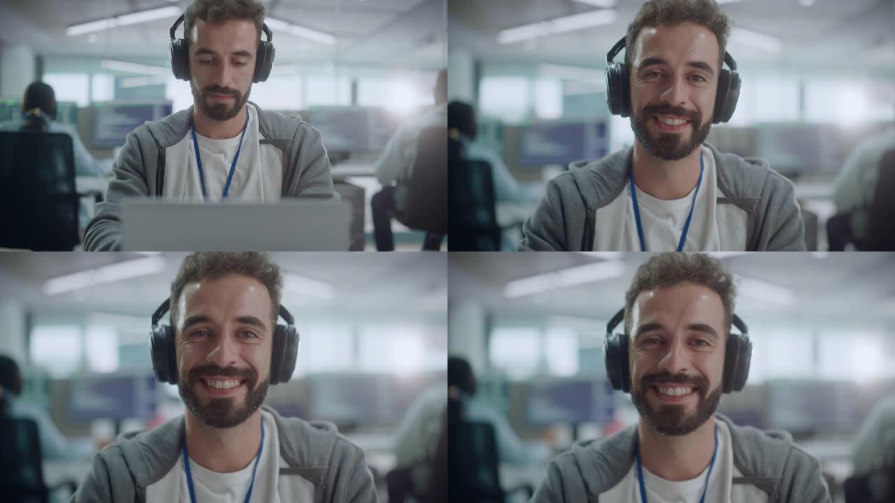 办公室: 快乐的IT程序员的肖像戴着耳机在台式计算机上工作，看着相机并微笑着。男软件工程师开发应用程
