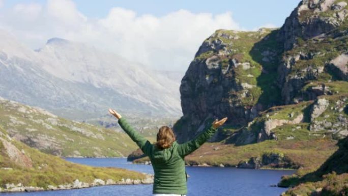 一个年轻女子张开双臂站着庆祝生活的后视图。无法识别的女人在冒险旅行度假中徒步穿越大自然时欣赏山景