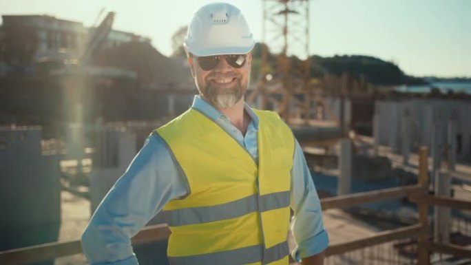 在阳光明媚的晴天，成功的大胡子土木工程师-戴墨镜的建筑师在建筑工地的镜头前微笑。男人戴着安全帽，衬衫