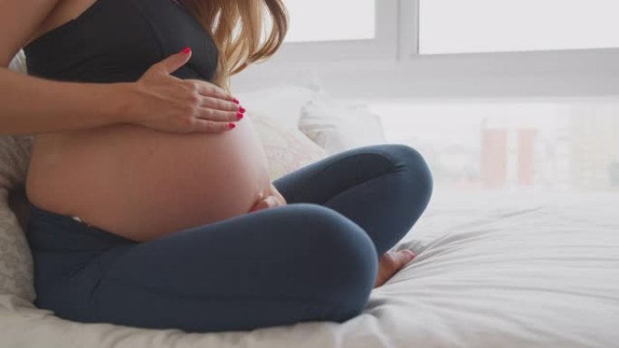 孕妇穿着健身服在家坐在床上摸肚子的特写
